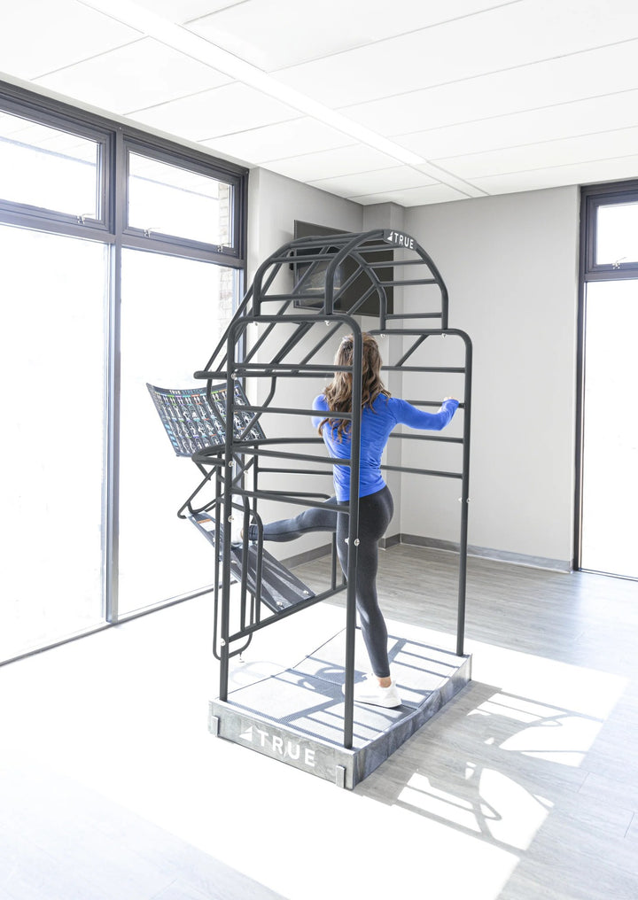 TRUE Stretch cage in use – woman doing a leg stretch against the frame in a bright gym